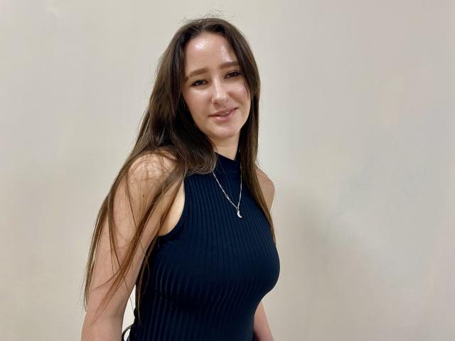 Omella has long brown hair, wearing a sleeveless black top, standing against a plain white background. She's smiling subtly, with a silver necklace, exuding a sexy, confident vibe.