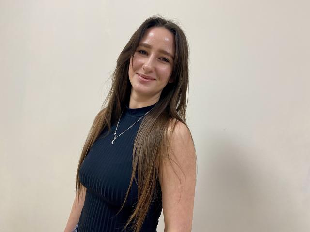 Photograph of Omella with long brown hair, wearing a sleeveless black top, smiling slightly, standing against a white background. She has a slight tilt, looking relaxed, with visible light reflection on her face.