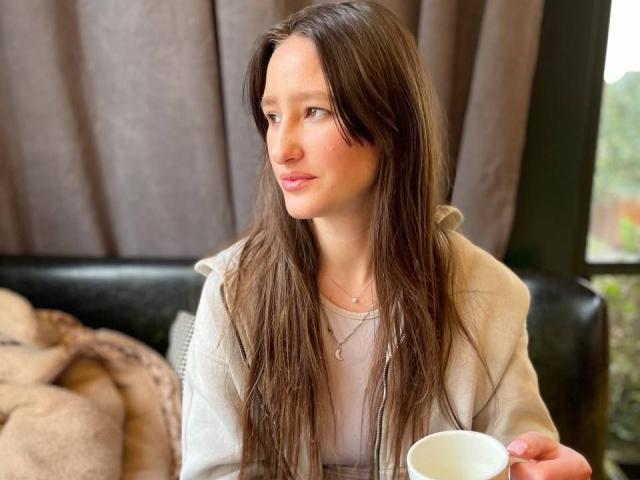 Photograph of Omella, a sexy woman with long brown hair, wearing a white coat, holding a white cup, looking to the side with a slightly aroused expression, sitting in a dimly lit room.