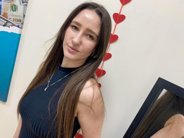 Omella with long brown hair, wearing a black sleeveless top, standing against a white wall with red heart decorations. Her gaze is direct, hinting at sexual tension. Mirror reflects her face.