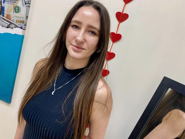 Omella mit langen, braunen Haaren, die ein schwarzes Top ohne Ärmel trägt, lächelt leicht und steht vor einer weißen Wand mit roten Akzenten. Herzdekorationen und ein teilweise sichtbares blaues Gemälde. Kette mit einem kleinen Anhänger.