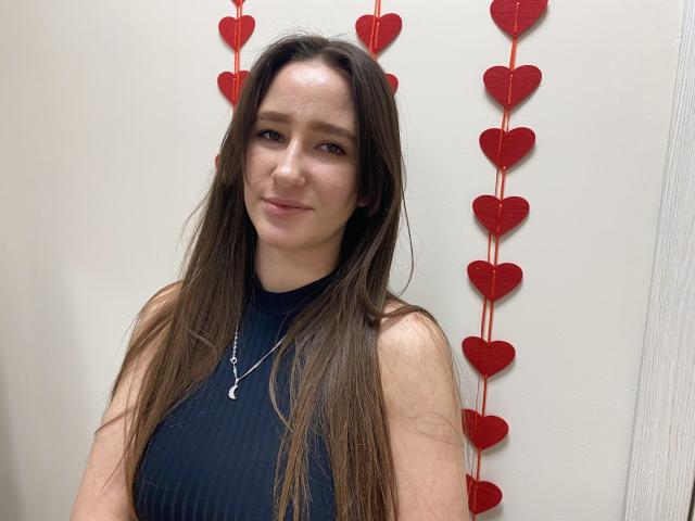 Omella, a brunette woman with long hair, wearing a black sleeveless top, stands against a white wall with red heart-shaped decorations. She has a slight smile, conveying subtle sexual tension.