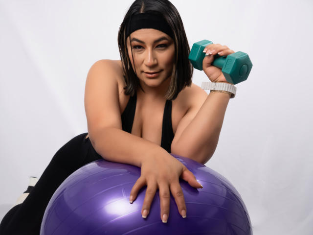 EmaFoxy, con el cabello negro y húmedo, se inclina sobre una pelota de ejercicios morada brillante, sosteniendo una mancuerna color turquesa, y lleva puesto un top negro. Pulsera blanca en la muñeca, mirando seductoramente a la cámara..