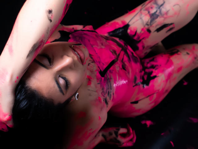 MiluSky, allongée sur le dos, recouverte de peinture rose vif et noire, les yeux à moitié fermés, la main sur la tête, peau brillante, regard perçant. tension sexuelle visible et intense, provocateur, fond sombre, gros plan..