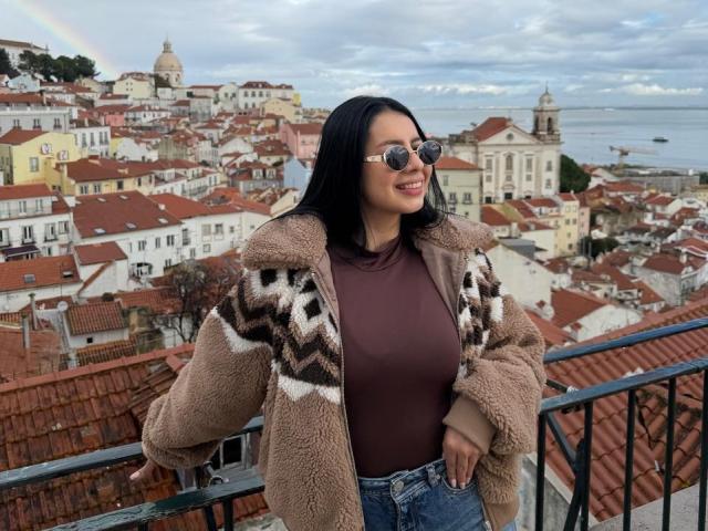 IsabellaJames with dark hair, wearing sunglasses, brown top, and patterned brown and white coat, leans on a railing with a view of a red-roofed, hilly European town in the background.