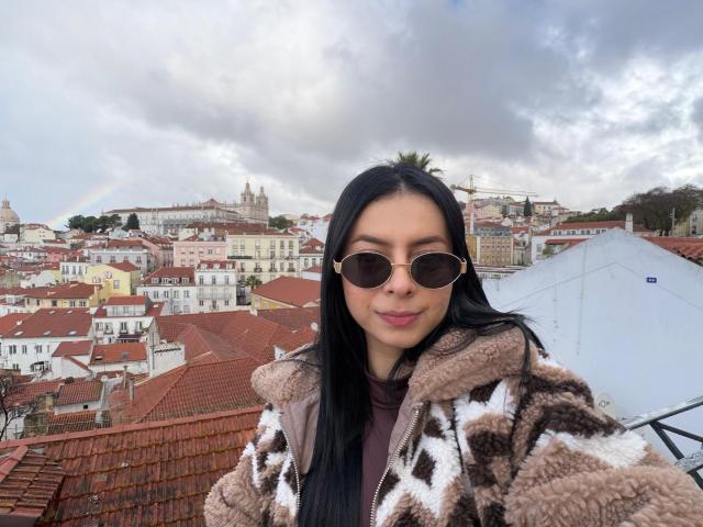 IsabellaJames with long black hair, wearing round sunglasses and a brown and white patterned fur coat, stands in front of a cityscape with red rooftops and cloudy sky. Fucking urban vibe.