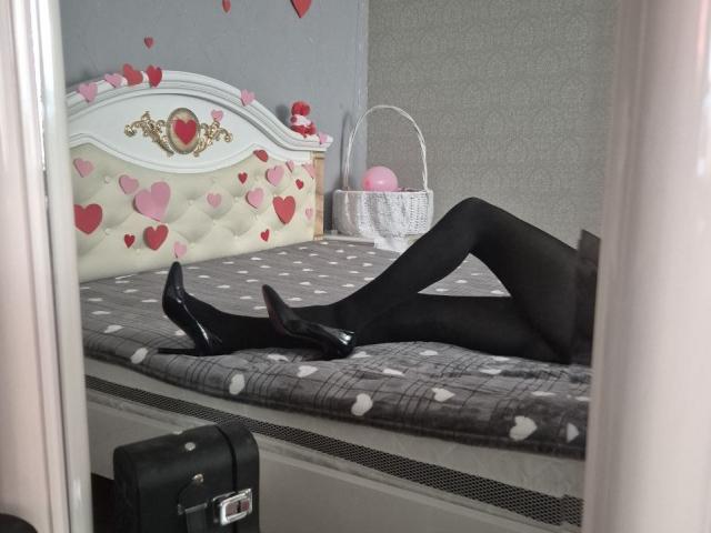 Anshante ligt op een bed, met gekruiste benen, en draagt zwarte panty's en zwarte hakken, met een wit hoofdeinde versierd met rode details. Hartstickers. Een rieten mand met roze ballen staat in de buurt..