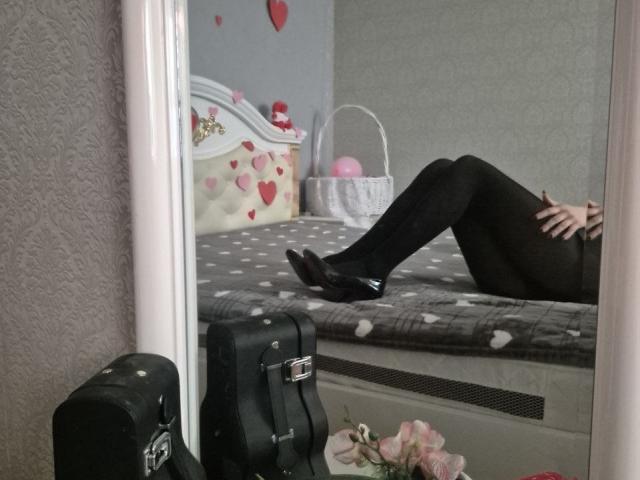Anshante reclines on a grey bed with white polka dots, legs crossed, wearing black pants and shoes. Mirror reflection shows a lit vertical light, suitcase, and pink flowers. Sexual tension.