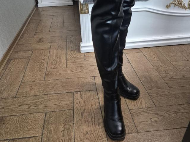 As botas de couro preto da Anshante, com solas grossas, estão sobre um piso de madeira com padrão de espinha de peixe. As calças pretas de Anshante estão levemente amassadas no joelho, projetando uma sombra no chão..