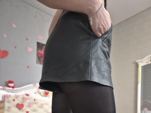 Close-up foto van Anshante, die een zwart leren top zonder mouwen en zwarte doorschijnende broek draagt, met haar hand in haar zak, zilver. armband, lichte grijze muur met rode hartstickers, zacht natuurlijk licht.