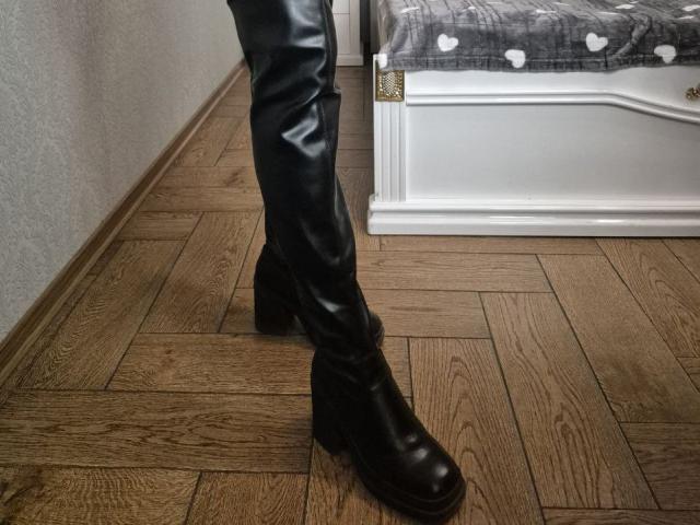 Anshante's lower legs and black leather boots stand on a wooden floor beside a white, illuminated box with a textured black top. Sexual tension from the poised, slightly provocative stance.