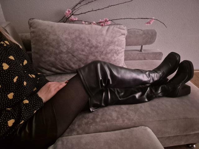 Anshante está deitada em um sofá cinza, com as pernas cruzadas nos tornozelos, vestindo botas de couro pretas e collant pretas. Ramo de cerejeira rosa como decoração ao fundo. O foco está em sua blusa preta com bolinhas..