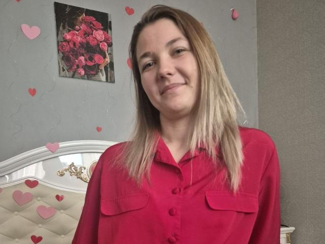 Anshante met recht blond haar, die een rood overhemd met knopen draagt en lichtjes glimlacht. Achtergrond: grijze muur, roze hartstickers, wit gestoffeerd hoofdeinde, schilderij van een roze roos. Ontspannen, sexy uitstraling.