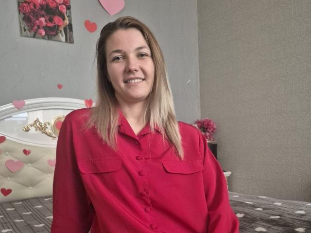 Anshante, con i capelli biondi lisci, indossa una camicia rossa con bottoni e si trova in una camera da letto. Cuori rossi e una parete rosa si trovano sopra un letto con una testiera imbottita. Tensione sessuale evidente..