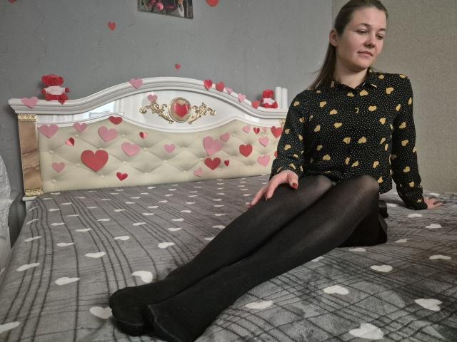 Anshante sitzt auf einem Bett mit einer grauen Decke mit Muster, trägt ein schwarzes gepunktetes Hemd und schwarze Leggings, die Beine sind gespreizt. Kopf leicht geneigt, wirkt entspannt. Kopfstütze im Hintergrund mit Herzmuster..