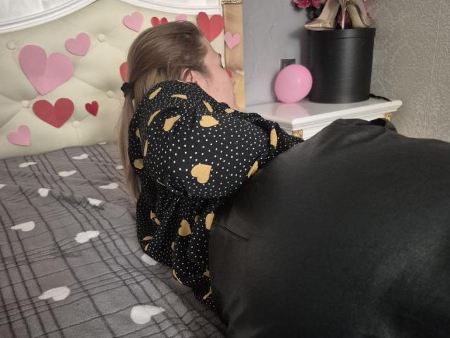 Anshante lies face-down on a bed with a plaid blanket, wearing a black polka-dot shirt with yellow stars, and black pants. Pink hearts and a pink balloon in the background.