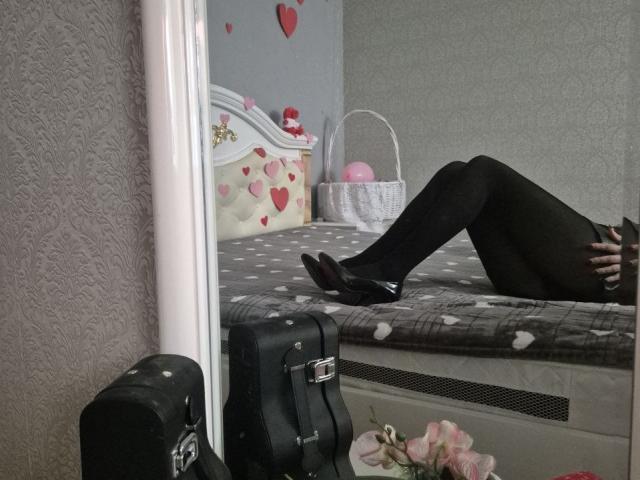 Anshante lag auf einem grauen Bett mit Herzmuster, die Beine hochgelegt, und trug schwarze Strumpfhosen und schwarze Schuhe. Ein schwarzer Koffer, ein weißer Korb mit rosa Herzen und rosa Blumen befinden sich in der Nähe. Verdammt heiß..