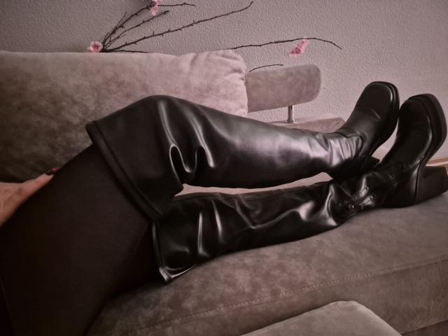 Anshante lies on a grey sofa, legs crossed at the ankles, wearing shiny black thigh-high boots. Pink cherry blossoms in the background. Sexual tension, legs prominent, boots glistening.