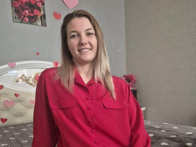 Anshante, com cabelo loiro reto e na altura dos ombros, vestindo uma camisa vermelha de botões, está em pé em um quarto com uma cama. Decorado com corações brancos e vermelhos. Ela está sorrindo, transmitindo uma aura sensual..