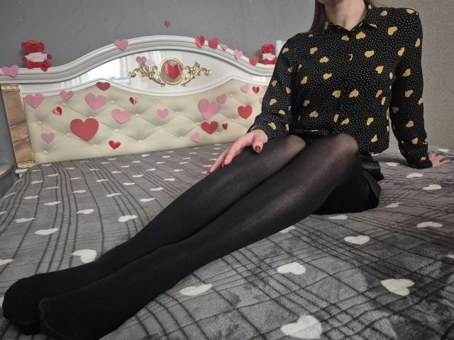 Anshante sits on a bed, legs extended, wearing a black polka-dot shirt with gold hearts and black pantyhose. The bed has a heart-patterned headboard and a gray plaid blanket.