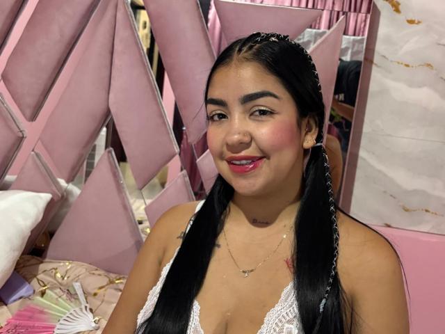 Natacha Luna, con cabello negro largo, rubor rosado y lápiz labial rojo, sonriendo a la cámara. Usando un sujetador de encaje blanco, rodeado de tela rosa en forma de triángulo. Increíblemente atractiva, sexy y segura de sí misma..