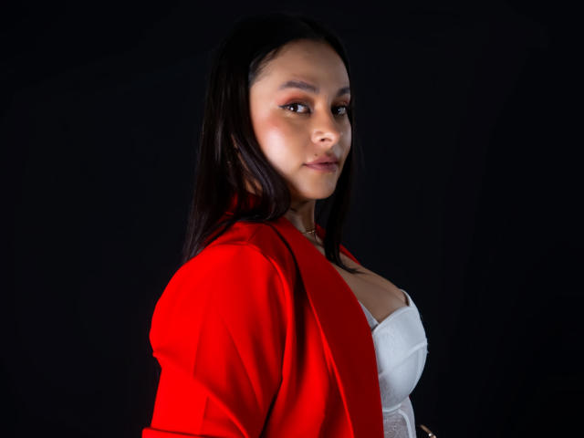 Photograph of LunaSoler, side profile, black hair, red jacket, white bra, black background, slight smile, direct gaze, highlighted cheek, shadows on face, light source from front, sensual, confident pose.