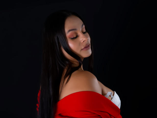 LunaSoler, met lang zwart haar, draagt een rode top met een open schouder, waarbij witte beugels van haar bh zichtbaar zijn. Ze kijkt met gesloten ogen naar beneden, met een verleidelijke, sensuele uitdrukking. De donkere achtergrond versterkt haar aantrekkingskracht..