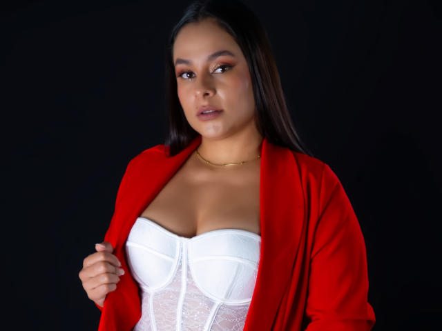 LunaSoler, with dark straight hair, gazes forward. She wears a red cardigan over a white lace corset, highlighting large breasts. Her expression is serious, with subtle makeup and a gold necklace.