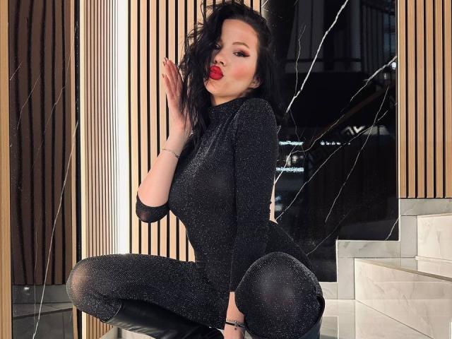 DeliciousChrissy si accovaccia in modo seducente, indossando una tuta nera scintillante e dei leggings neri. Ha i capelli scuri e mossi, un rossetto rosso acceso e un'espressione seducente e attraente. La sua mano tocca il suo viso
.