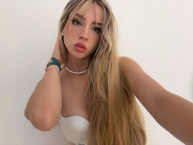 Amelia Collins hat langes, blondes Haar, eine Ponyfrisur und volle, rote Lippen. Sie trägt ein weißes Top ohne Träger, eine Perlenkette und ein grünes Armband. Sie blickt direkt in die Kamera und ihr Blick ist verführerisch..