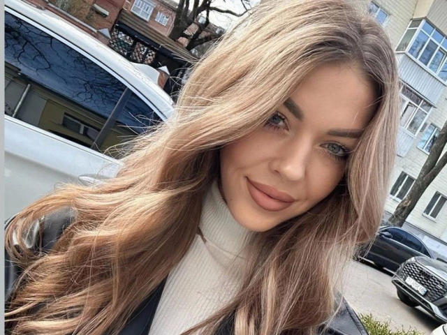 Nahaufnahme von LizaSweety, attraktiv mit langen, welligen braunen Haaren, blauen Augen und vollen Lippen, die ein weißes Rollkragenpullover trägt und steht. Neben einem weißen Auto, Hintergrund einer Straße mit Gebäuden und Bäumen. Verdammt heiß..