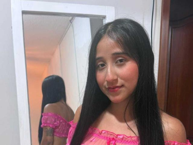 AnnieBlakke with long black hair, pink off-shoulder top, smiling at camera, reflection in mirror shows back, slight cleavage, bedroom setting, soft light, casual, sexy, attractive, confident pose.