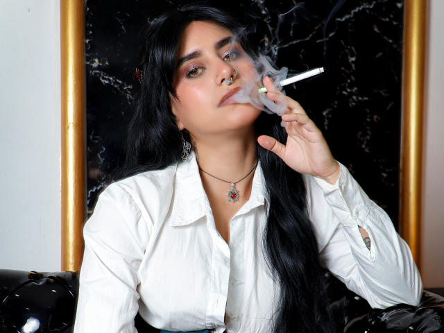 SamanthaGio, con su largo cabello negro, fuma un cigarrillo, exhalando humo. Vestida con una camisa blanca y un collar, está mirando a la cámara con una expresión ligeramente seductora. Fondo oscuro.