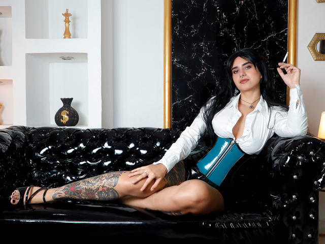 SamanthaGio, une femme aux formes généreuses avec des cheveux foncés, est assise sur un canapé noir texturé. Elle porte une chemise blanche, un corset bleu et une jupe noire, et on peut voir une grande tatouage sur sa jambe. Extrêmement sexy..