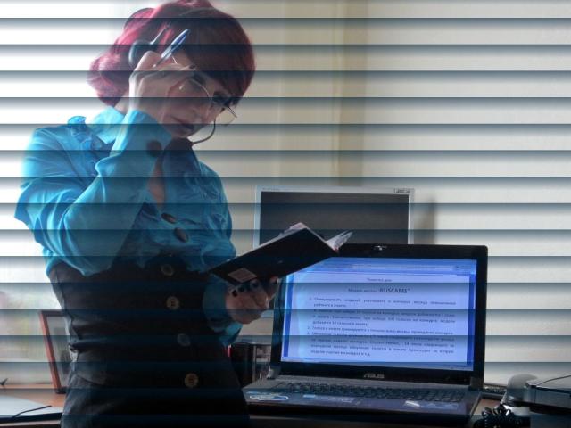 FetishDreams4u, uma mulher atraente com cabelo ruivo e óculos, vestindo uma camisa azul, está em frente a um laptop com a tela exibindo texto. Segurando um livro, concentrando-se nele. Persianas ao fundo..