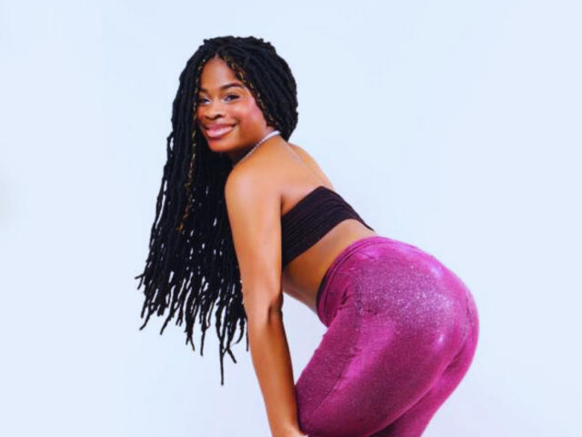 TamaraLubezki bends forward, smiling, with long black dreadlocks. She wears a black tube top and tight, sparkly pink pants, emphasizing her curvy butt. White background, casual pose, sexy vibe.
