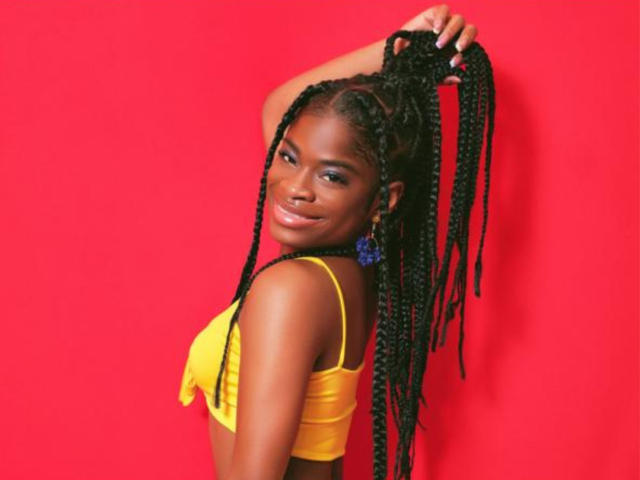 TamaraLubezki with dark skin and long black braids, wearing a yellow crop top, smiling at the camera, hand raised, touching her hair, against a bright red background, looking sexy.