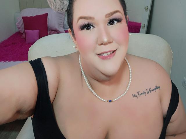Photograph of a plump woman with fair skin, black hair, and blue eyes, wearing a black strapless top and pearl necklace with "My Treasure is You" tattoo. Cocky smile, pink blush.