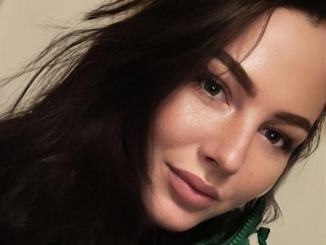 Fotografía de primer plano de AnnieQx, con el rostro ligeramente inclinado hacia la derecha, cabello castaño oscuro, luz suave en la piel, una sonrisa sutil, color verde. ojos, maquillaje ligero, sombras suaves, expresión íntima y relajada..