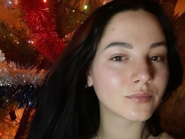 Close-up of AnnieQx with dark hair, fair skin, and subtle makeup, gazing directly at the camera. Red and silver Christmas lights in the background create a warm, festive atmosphere.