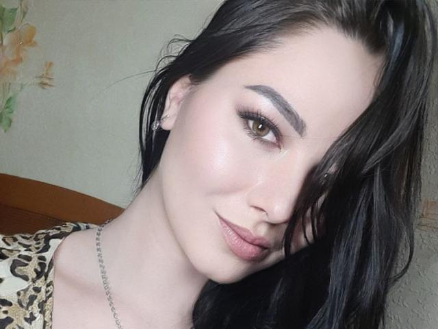 Close-up of AnnieQx, pale-skinned woman with dark hair, green eyes, and full lips, wearing leopard print top, silver chain necklace, tilting head slightly to the right, sexy, seductive expression.