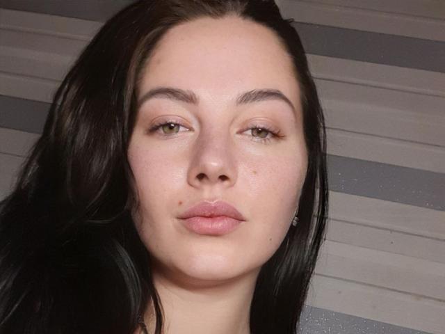 Close-up of AnnieQx, a pale-skinned woman with long black hair, full lips, and brown eyes. Her expression is neutral, slightly seductive. Stripes in the background. Skin has a slight sheen.