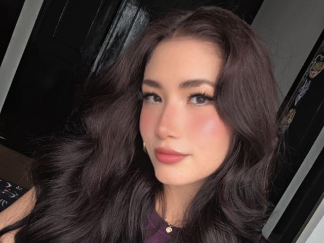 Gros plan de StarDustt, une femme séduisante avec de longs cheveux noirs ondulés, une peau claire et des yeux foncés, portant un haut violet, sobre. du maquillage, et un collier en or, avec un sourire légèrement séduisant..