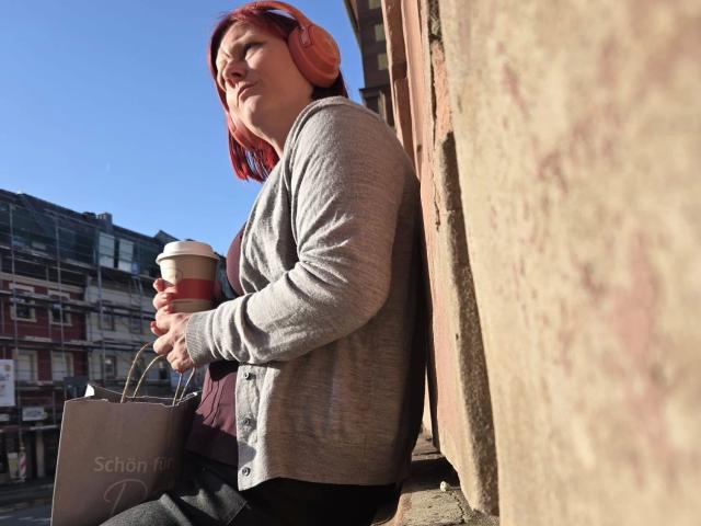 Ella-Bizarr-hot, zittend tegen een gebouw, met een koffiekop en een bruine boodschappentas, gekleed in een grijze cardigan en rode hoofdtelefoon, zonlicht dat haar rode haar accentueert, stedelijke straat als achtergrond..