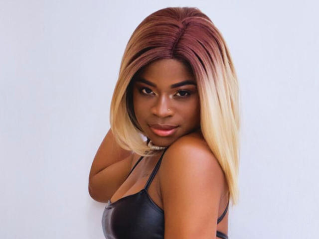 Photograph of TamaraLubezki, dark-skinned woman with shoulder-length, ombre hair transitioning from dark brown to blonde. She's wearing a black leather top, looking directly at the camera with a seductive gaze.