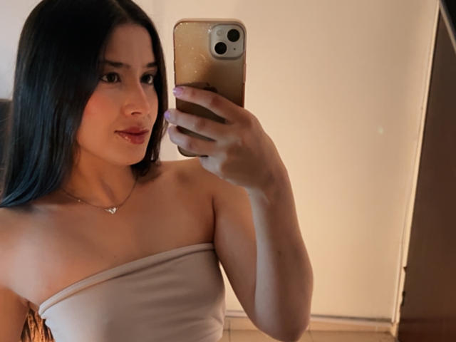 EmmaLombardi prend un selfie devant un miroir, tenant un téléphone doré, et porte un haut blanc sans bretelles. Elle a de longs cheveux noirs, une peau brun clair et un sourire discret. Le fond est légèrement éclairé..