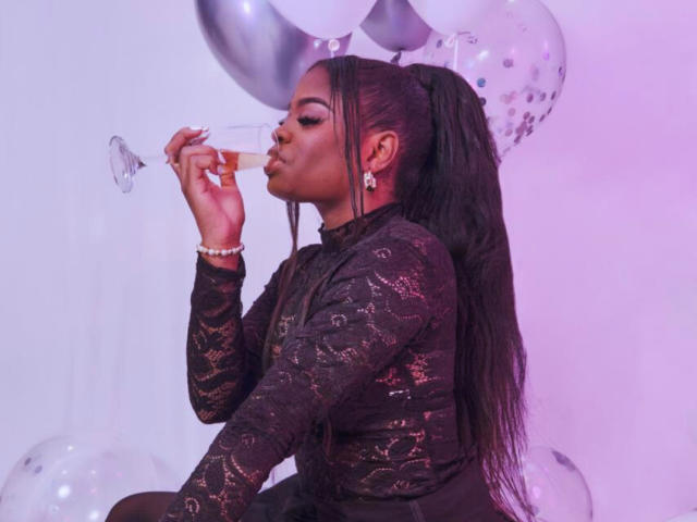 TamaraLubezki, a Black woman with long, straight black hair in a high ponytail, blows on a bubblegum bubble while wearing a black lace long-sleeve top. Purple and white balloons surround her.