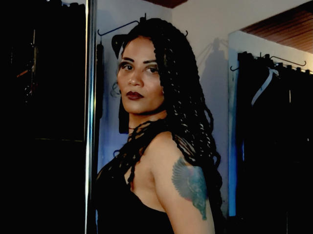 Photograph of DivaMarie, a curvy woman with long black dreadlocks, dark lipstick, and a tattoo on her upper arm, wearing a black sleeveless top, standing in front of dark clothes. Fucking sexy.