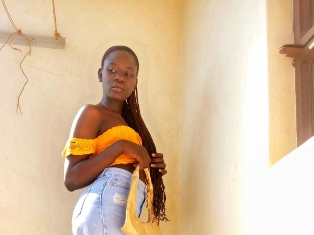 ShanyShar se tient dans une pièce baignée de soleil, vêtue d'un haut jaune à une épaule et d'un jean taille haute. De longues tresses descendent le long de son dos. Une lumière vive projette de fortes ombres sur sa peau foncée. Incroyablement sexy.