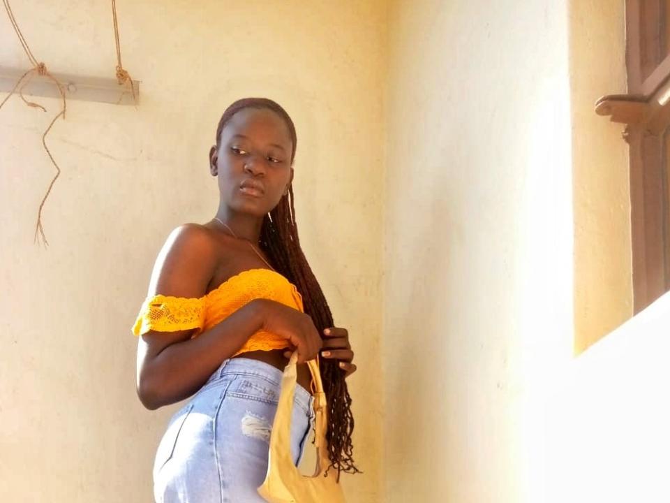 ShanyShar está de pie en una habitación iluminada por el sol, vistiendo una blusa amarilla con un hombro descubierto y jeans de cintura alta. Las largas trenzas le caen por la espalda. Una luz brillante proyecta sombras marcadas sobre su piel oscura. Increíblemente atractivo..